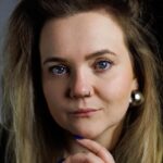 młoda kobieta, ciemna blondynka, patrzy się prosto w obiektyw, lekko się uśmiechając, ma rozpuszczone włosy, widoczne jest jedno ucho z srebrnym kolczykiem w kształcie kuli, lekko jednym palcem podpiera podbródek. Jest to Joanna Piecyk