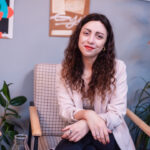 młoda uśmiechnięta kobieta w długich kręconych ciemnych włosach. Siedzi na retro fotelu z założoną nogą na nogę i skrzyżowanymi dłońmi ułożonymi na kolanie.