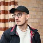 młody mężczyzna w czarnej czapce z daszkiem z lekkim zarostem, w rozpiętej czarnej bluzie sportowej i jasnym tiszercie, patrzy w lewą stronę. Jest to Mateusz Demski