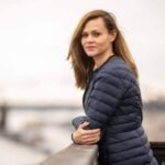 młoda kobieta z ciemnymi włosami do ramion, stoi bokiem z głową zwróconą w stronę obiektywu, jest na zewnątrz i opiera się o barierkę