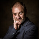 łysiejący mężczyzna w średnim wieku, podpiera podbródek jedną ręką, lekko się uśmiecha