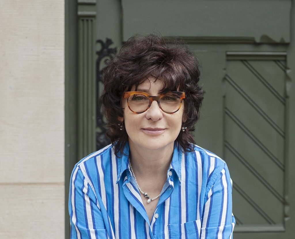 Kobieta, brunetka w krótkich kręconych włosach i okularach w pasiastej koszuli, siedzi na schodach. Jest to Joanna Cichocka-Gula