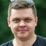 mężczyzna, brunet, lekko uśmiechnięty w ciemnym tiszercie z okularami przeciwsłonecznymi założonymi na czubku głowy