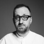 czarnobiały portret mężczyzny w białej koszuli. mężczyzna ma zarost i okulary korekcyjne. Jest to Michał Nogaś